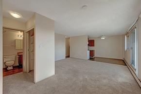 Edmonton apartment for rent - A spacious open living area here at Cedar Gardens with soft carpeting and an easy flow into the kitchen space, featuring large sliding glass doors with wide windows for natural light, a long baseboard heater, and nearby built-in shelving and bathroom access for added everyday convenience.