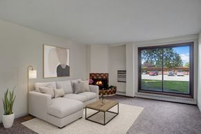 Saskatoon apartment for rent - A cozy living room here at Edgemont Heights featuring a soft light gray sectional with neutral accent pillows, a warm wood coffee table on a textured area rug, and a glowing fireplace, complemented by simple wall art, greenery, and a large picture window with peaceful outdoor views.