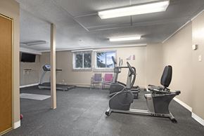Saskatoon apartment for rent - A bright fitness room here at Edgemont Heights featuring cardio equipment like a treadmill, elliptical, and stationary bike, plus open floor space for stretching or strength training, all in a clean, simple setting with windows for natural light.