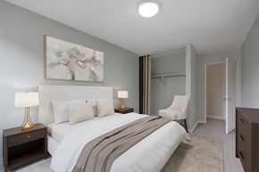 St. Albert apartment for rent - A cozy bedroom here at Gateway Gardens featuring a comfortable bed with crisp white bedding and neutral pillows, paired with matching nightstands and warm table lamps, plus simple wall art, an accent chair, and an open closet for easy storage.