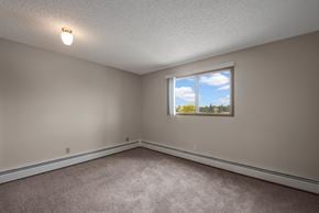 St. Albert apartment for rent - A bright, empty bedroom here at Gateway Square with soft beige walls, plush carpet flooring, and a large window with vertical blinds letting in natural light and peaceful outdoor views.