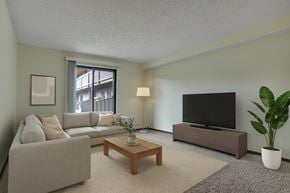 Saskatoon apartment for rent - A bright living room here at Kingsmere with soft green walls, plush carpet, a light neutral sectional sofa, a wooden coffee table, and a media console, with natural light coming in through a sliding glass door for a calm, inviting feel.