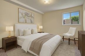 Saskatoon apartment for rent - A softly staged bedroom here at Kingsmere with neutral walls, plush carpet, a comfortable bed with layered bedding, warm accent lighting, and a window bringing in natural light for a calm, inviting atmosphere.