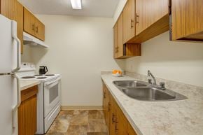 Edmonton apartment for rent - A clean, functional kitchen here at Lakewood Heights with warm wood cabinets, light countertops, a stainless steel double sink, white appliances, and efficient galley-style layout with bright overhead lighting.