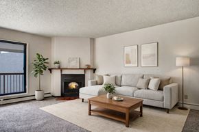 Regina apartment for rent - A warm, inviting living room here at Madison Park featuring a neutral sectional sofa with textured throw pillows, a wooden coffee table, soft area rug, and a cozy fireplace with a simple mantel, complemented by natural light from a sliding glass door leading to a private balcony.