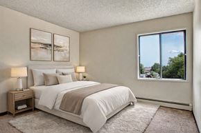 Regina apartment for rent - A bright, calming bedroom here at Madison Park featuring a neatly made bed with white bedding, layered neutral pillows, and a soft throw, paired with matching nightstands and warm table lamps, framed by minimalist wall art, plush carpet, and a large window offering natural light and neighborhood views.
