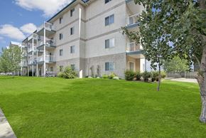 Edmonton apartment for rent - A landscaped apartment building exterior here at Madison Park featuring a multi-story beige building with private balconies and large windows. A wide green lawn stretches across the foreground, bordered by mature trees, shrubs, and garden beds, creating an open, park-like setting under a bright blue sky.