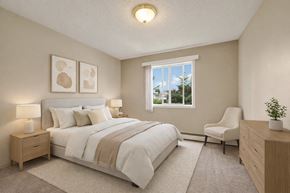 Edmonton apartment for rent - A calm, inviting bedroom here at Madison Park with beige carpet, soft neutral walls, and a large window bringing in natural light and leafy outdoor views. A neatly made bed with white bedding and layered neutral pillows sits between matching nightstands with lamps, complemented by abstract wall art, a cozy accent chair, and a light wood dresser.
