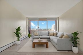Saskatoon apartment for rent - A bright, inviting living room here at Madison Park with soft neutral walls, beige carpet, and large sliding glass doors that fill the space with natural light and city views. A light gray sectional sofa with neutral pillows sits on an area rug, paired with a wooden coffee table and potted plants, creating a comfortable, modern setting.