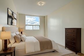 Saskatoon apartment for rent - A bright, comfortable bedroom here at Madison Park with soft neutral walls, plush carpet, and a large window that brings in abundant natural light and open sky views. A neatly made bed with layered white and beige bedding sits beside matching bedside tables with lamps, complemented by a dark wood dresser and simple wall art.