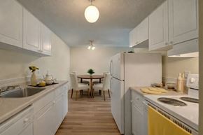 Edmonton apartment for rent - A bright, functional kitchen here at Park Place South featuring crisp white cabinetry, light countertops, and clean appliances, with warm wood-style flooring that opens into a cozy dining area with a round table and upholstered chairs, creating an inviting space for everyday cooking and casual meals.