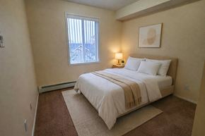 Calgary apartment for rent - A cozy bedroom here at Pointe at Applewood with soft carpet, neutral walls, and a large window with vertical blinds. A full bed with white bedding and a bedside table creates a warm and inviting atmosphere.
