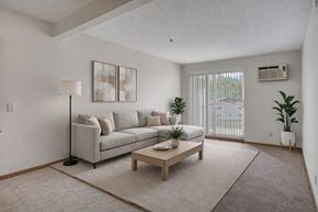 Winnipeg apartment for rent - A bright living room here at Quail Ridge with soft carpet flooring, a light-gray sectional sofa, and a simple wooden coffee table on a neutral area rug. A large window with vertical blinds brings in natural light, creating a clean and relaxing vibe.