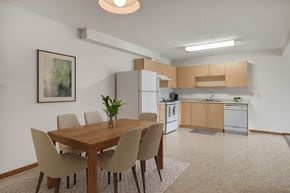 Winnipeg apartment for rent - A bright dining area here at Quail Ridge with a wooden table, four upholstered chairs, and simple wall art for a clean, welcoming feel. A spacious kitchen sits just beyond with light wood cabinets, white appliances, and a neutral tile floor.