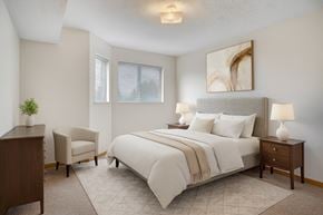 Winnipeg apartment for rent - A bright bedroom here at Quail Ridge with soft carpet, neutral walls, and a cozy bed dressed in crisp white bedding and warm accents. A pair of nightstands with matching lamps and a cushioned chair create a calm, inviting space with gentle natural light from the windows.