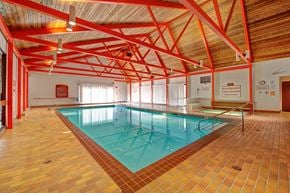 Grande Prairie apartment for rent - A spacious indoor swimming pool here at Ranchlands showcasing a rectangular lap pool with clear water, tiled pool deck, metal handrails, and safety signage, set beneath a vaulted wood ceiling with exposed red beams and ample overhead lighting that creates an open, recreational atmosphere.