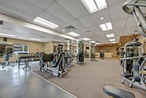 Grande Prairie apartment for rent - A well-equipped fitness center here at Ranchlands featuring a spacious layout with strength training machines, free weights, and cardio equipment arranged across carpeted and rubber flooring, complemented by mirrored walls, overhead lighting, and neutral finishes that support a comfortable workout environment.
