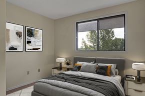 Grande Prairie apartment for rent - A cozy modern bedroom here at Ranchlands featuring a queen bed with layered neutral bedding and accent pillows, a soft upholstered headboard, matching nightstands with table lamps, abstract wall art, plush carpet flooring, and a large window bringing in natural light and leafy outdoor views.