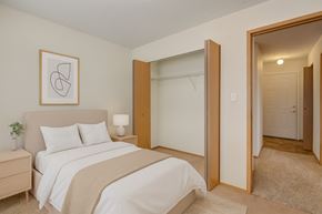 White Court apartment for rent - A cozy, neutral bedroom here at Sol Terrace featuring a neatly made bed with soft white bedding and a textured throw, matching bedside tables with warm wood tones, an open closet with sliding wood doors, and a hallway view that highlights comfortable carpeting and clean, simple finishes throughout the space.