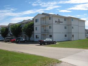 White Court apartment for rent - A landscaped exterior view here at Sol Terrace showcasing a three-story apartment building with light-colored siding, private balconies, surrounding green lawn areas, mature trees, and convenient on-site parking along a paved drive under a clear blue sky.