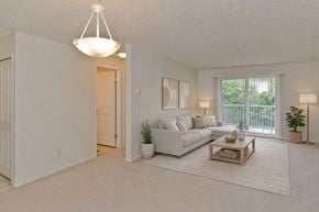 Calgary apartment for rent - A spacious, neutral living room here at Somerset Pointe featuring a light gray sofa with coordinating pillows, a wooden coffee table on a textured area rug, soft overhead lighting, and a sliding glass door with vertical blinds that opens to greenery, creating a calm and comfortable gathering space.