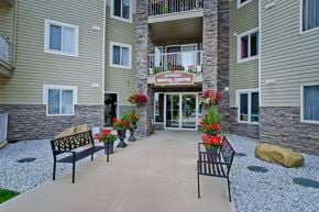 Calgary apartment for rent - A welcoming exterior entrance here at Somerset Pointe featuring the rental centre doorway framed by stone accents, beige siding, and colorful flower planters, with benches along the concrete walkway and neatly landscaped gravel beds creating an inviting apartment community arrival.