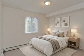 Calgary apartment for rent - A serene bedroom here at Somerset Pointe featuring a plush neutral bed with layered white bedding and soft accent pillows, matching nightstands with warm table lamps, light carpeting, minimalist wall art above the headboard, and a window with vertical blinds that brings in gentle natural light.
