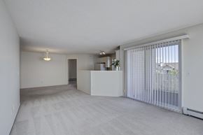 Calgary apartment for rent - A bright open living room here at Somerset Pointe featuring soft neutral carpet, light gray walls, and a spacious layout that connects to a partial kitchen pass-through, highlighted by modern ceiling lighting and a large sliding glass door with vertical blinds that opens to a private balcony and brings in abundant natural light.