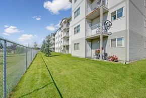 Stony Plain apartment for rent - A landscaped exterior view here at Sonora showcasing a well-maintained green lawn alongside a multi-story apartment building with light-colored siding, private balconies on each level, patio seating with potted flowers, mature trees, and a fenced perimeter under a bright blue sky.