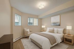 Stony Plain apartment for rent - A cozy, light-filled bedroom here at Sonora featuring a queen-size bed with crisp white bedding, a soft neutral throw, matching bedside tables with warm lamps, plush carpet flooring, a comfortable accent chair, and windows with blinds that bring in natural light for a calm, inviting retreat.