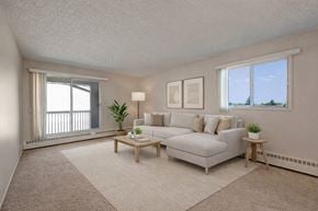 Edmonton apartment for rent - A bright living room here at Stirling Place featuring a spacious layout with soft neutral carpeting, a light-colored sectional sofa with coordinating pillows, a wooden coffee table and side table, simple abstract wall art, large windows bringing in natural light, and sliding glass doors leading to a private balcony.
