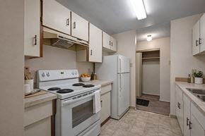 Edmonton apartment for rent - A clean galley-style kitchen here at Stirling Place featuring white cabinetry with dark hardware, light countertops, a white electric stove and refrigerator, a double-basin sink, vinyl flooring, overhead lighting, and an adjacent hallway with a storage closet creating a functional and efficient layout.
