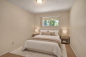 Edmonton apartment for rent - A cozy bedroom here at Stirling Place featuring a queen-sized bed with neutral bedding and layered pillows, matching nightstands with soft table lamps, plush carpet flooring, light beige walls, and a window above the headboard that brings in natural light and a peaceful outdoor view.