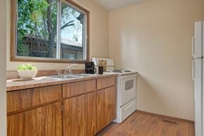 Saskatoon apartment for rent - A bright functional kitchen here at Ventana featuring warm wood cabinetry, a laminate countertop with a double sink beneath a large window, a white electric range and refrigerator, vinyl plank flooring, and simple countertop accents, with natural light highlighting the clean layout and practical workspace.