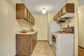 Saskatoon apartment for rent - A warm compact kitchen here at Ventana showcasing rich wood cabinetry, a laminate countertop with integrated sink, white electric range and refrigerator, vinyl tile flooring, and simple staged accents, with overhead lighting and a streamlined galley layout that maximizes functionality and storage.