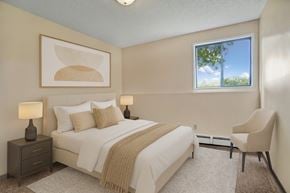 Saskatoon apartment for rent - A serene bedroom here at Ventana featuring a queen-sized bed with crisp white bedding, layered neutral pillows, and a soft beige throw, complemented by matching bedside tables with lamps, abstract wall art above the headboard, plush carpeting, a cozy accent chair, and a bright window bringing in natural light.