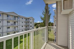 Winnipeg apartment for rent - A bright private balcony here at Villagio overlooking landscaped green lawns and neighboring apartment buildings, featuring white railing, covered outdoor space, and open sky views that provide a relaxing spot for fresh air, outdoor seating, and enjoying the surrounding community scenery.