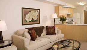Saskatoon apartment for rent - A warm and inviting living room here at Wellington Manor with a plush cream sofa, a round glass coffee table, and soft accent lighting. A floral wall art piece and patterned throw pillows add a cozy touch to the space. A bright open kitchen is visible in the background with light wood cabinets, a breakfast bar, and white appliances for a practical layout.