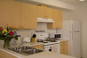 Saskatoon apartment for rent - A bright kitchen here at Wellington Manor with light wood cabinets, clean white countertops, and a double stainless steel sink. A white stove with a vent hood, a microwave, and a full-size refrigerator provide a practical cooking setup. A wide breakfast bar counter adds extra prep space and a convenient spot for casual dining.