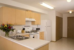 Saskatoon apartment for rent - A spacious kitchen here at Wellington Manor with light wood cabinets, white countertops, and bright overhead lighting. A double stainless steel sink sits along a wide breakfast bar that offers extra counter space for cooking or casual dining. A full appliance set includes a white stove with vent hood, a microwave, and a full-size refrigerator for everyday convenience.