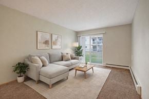 Lethbridge apartment for rent - A bright living room here at Westhaven Estates with soft carpet flooring, a light gray sectional sofa, and a neutral area rug for a cozy feel. A sliding glass door lets in natural light and opens to a grassy outdoor view. A simple wooden coffee table, minimal wall art, and greenery accents add a clean and relaxing vibe.