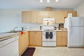 Lethbridge apartment for rent - A bright kitchen here at Westhaven Estates with light wood cabinets, white countertops, and clean tile flooring. A full appliance set includes a white refrigerator, stove with vent hood, and a built-in dishwasher for everyday convenience. A double sink and generous counter space create a practical layout with a warm, welcoming feel.