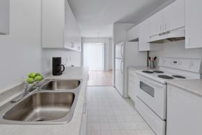 Dawson Creek apartment for rent - A clean galley-style kitchen here at Willowview Terrace with white cabinets, a double stainless-steel sink, and bright countertops for a simple modern look. A white refrigerator and electric stove sit along the right side, with light flooring and a dining area visible near the sliding glass doors at the back.