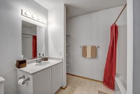 Dawson Creek apartment for rent - A clean bathroom vanity area here at Willowview Terrace with a white sink countertop, a large wall mirror, and bright overhead lighting. Open shelving provides extra storage space, while a towel bar and a simple tub-and-shower combo with a red shower curtain complete the practical layout.