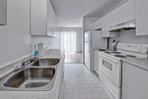 Dawson Creek apartment for rent - A bright galley-style kitchen here at Willowview Terrace with white cabinets, light countertops, and a double stainless steel sink. White appliances including a stove, range hood, and refrigerator line the space, with durable tile flooring that leads into the dining area with a sliding glass door for natural light.