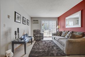 Saskatoon apartment for rent - A cozy living room here at Windsor Terrace with a soft gray sofa, patterned accent chair, and a dark area rug that anchors the space. A bold red accent wall with a large mirror and warm table lamp adds character, while framed wall art and a console table provide simple decorative touches. Sliding glass doors bring in natural light and open to a patio area outside.