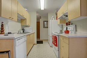 Saskatoon apartment for rent - A bright galley-style kitchen here at Windsor Terrace with light wood cabinets, white countertops, and easy-to-clean tile flooring. This space includes a double sink, dishwasher, and a full set of white appliances, with overhead lighting that keeps the room feeling open and practical. Simple countertop decor and warm red accents add a welcoming touch.