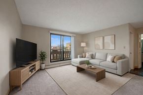 Edmonton apartment for rent - A bright living room here at Wyndham Crossing with a cozy light gray sectional sofa, neutral throw pillows, and simple wall art above the seating area. A large sliding glass door brings in natural light and opens to a private balcony, while a modern TV console, soft area rug, and small coffee table complete the clean and inviting space.