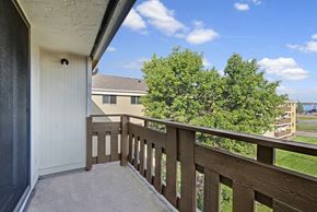 Edmonton apartment for rent - A private balcony view here at Wyndham Crossing with a concrete patio floor and a sturdy brown railing overlooking green trees and nearby buildings. The bright blue sky with soft clouds adds a fresh, open feel to the outdoor space.