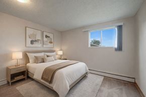 Edmonton apartment for rent - A bright, relaxing bedroom here at Wyndham Crossing with a cozy bed dressed in crisp white bedding, neutral accent pillows, and a soft beige throw blanket. Two matching nightstands with modern lamps sit on either side, while simple wall art and a large window bring in natural light above the plush carpet.